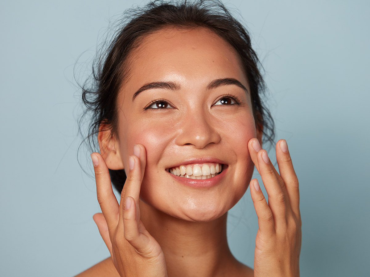 A woman in front of a light blue background, smiling and lightly touching her fingers to her red cheeks.
