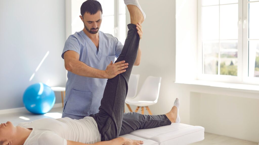 man in scrubs assists patient in a straight leg raise while the patient lays on an exam/therapy table