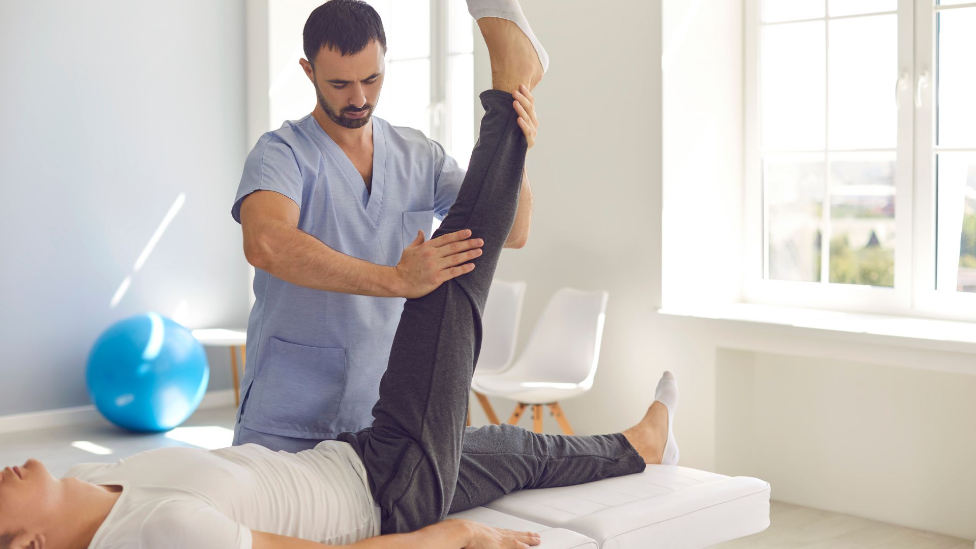 man in scrubs assists patient in a straight leg raise while the patient lays on an exam/therapy table
